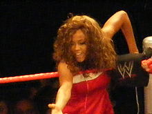 A curly haired woman stands on the apron of a wrestling ring with red ropes at the corner turnbuckle. The turnbuckle is black and has the red and white WWE logo on it. The woman is wearing a red dress with a whit trim across the top.