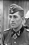 A man wearing a military uniform, side cap and neck order in the shape of a cross. His cap has an emblem in shape of a human skull and crossed bones.