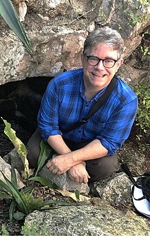 David Stuart wearing a blue checkered shirt and gray pants, squatting down amid rocks, grinning up at camera