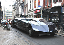 A dark-blue stretch-limousine-type vehicle in a narrow shopping street