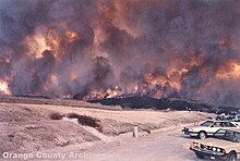 Cars parked askew near a beach to the right of a thin line of flame on the horizon, with translucent plumes of smoke filling the sky.