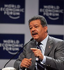 A portrait shot of a middle-aged man smiling somewhat and looking straight ahead. He has light brown skin, slightly African/Negroid facial features, curly dark hair. He is mustachioed and wears a suit and tie.