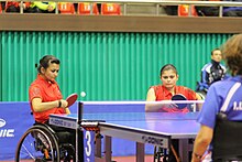 Zwei Frauen im Rollstuhl mit Tischtennisschlägern an einem blauen Tischtennistisch.