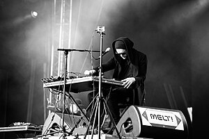 black-and-white image of Sohn wearing dark clothing, a tuque, and dark sunglasses, standing and operating a control board onstage