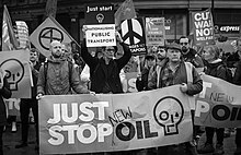 Black and white photo. There is a crowd of about twenty people. Most hold protest signs. Three people stand in the foreground. Two of them hold a banner with the Just Stop Oil logo; the banner reads "Just stop new oil" and the word "new" appears to have been added by hand. The other person in the foreground holds a sign with the Just Stop Oil logo. It reads "Just start nationalising public transport".