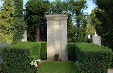 A tall stone column bearing the words "Gustav Mahler", surrounded by a low green hedge, with a floral bloom in the foreground