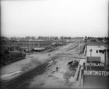 A few 2-story commercial brick buildings are on each side of the dirt street with puddles from rain. A horse-drawn wagon and a man are in the street.