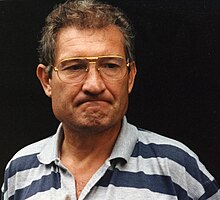 Photographie en buste d'un homme vêtu d'un polo rayé gris et bleu et portant des lunettes.