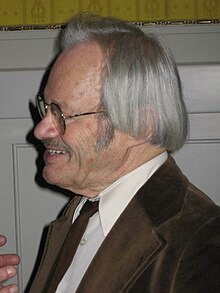 Side profile of a smiling, silver haired, older, white man, wearing glasses and a brown velvet suit jacket, white shirt and a brown tie.