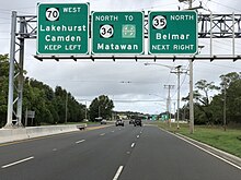 A six-lane divided highway approaching an intersection with a set of three green signs over the roadway. The left sign reads Route 70 west Lakehurst Camden keep left, the middle sign reads north Route 34 to Garden State Parkway Matawan, and the right sign reads Route 35 north Belmar next right.