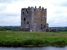 A massive, roofless four storey medieval stone tower sits amid grass and trees. On one side the stones are stained with orange lichen.