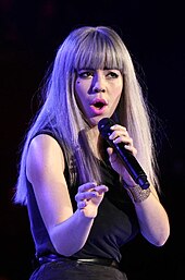 A young blonde woman with a black heart on her cheek singing into a microphone. She is wearing a black dress.