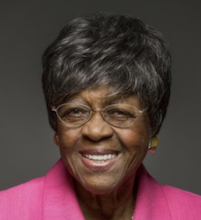 A headhsot of an older black woman who is smiling. She has short dark hair and is wearing a bright fuschia top