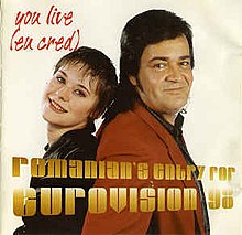 A shot of Olinescu and Adrian Romcescu standing side to side in front of a beige backdrop. Information on the song is superimposed on them in yellow and red letters.
