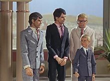Three men and a boy stand in a palatial setting. A desert landscape is visible from a balcony in the background. One man on the far left is grey-haired and wears a grey suit and tie, the man to the right of him dark-haired and in deep navy blue. Both men are orientated in the direction of the (blond-haired) boy, who is also formally attired in grey. The third man, also blond but wearing a cream-shaded suit, stands directly behind him.