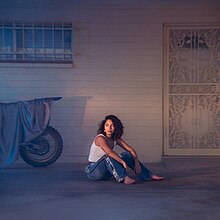 A photo of the artist sat on the ground in front of a house.