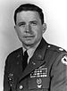 Head and shoulders of a white man with dark hair wearing a military jacket with a round patch on the upper sleeve, and oak leaf emblem atop the shoulder, and ribbon bars and pins on the left breast.