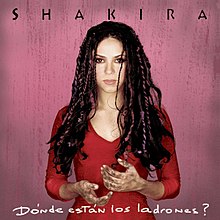 A brunette woman is in front of a pink background. She is wearing a red blouse and colorful braids, and her hands are covered in dirt. The word "Shakira" is above her, stylized in all capital letters, while the words "Dónde Están los Ladrones?" are placed beneath her.