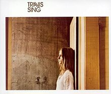 A woman with dark blonde hair, facing left, stands against a white door behind her. Her head is turned slightly toward the viewer. The background is a wooden wall. Surrounding this picture is a white border with a thicker top line. On this line, halfway between the left edge and center, is written "TRAVIS", and below it, "SING".