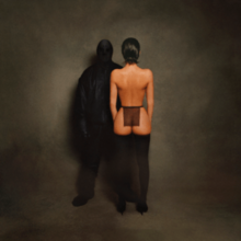 A photograph of West completely obscured by all-black clothing (including an opaque head covering) standing next to his wife, Bianca Censori, with her back turned, wearing stockings and a sheer black cloth around her waist