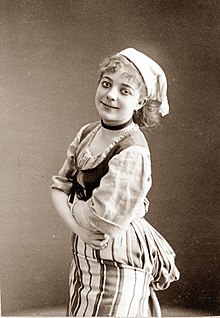 Photo of young white woman in 17th-18th century costume with a kerchief around her mid-length hair, facing to the left, crossing her arms and grinning towards the camera
