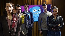 Standing in a line: a woman wearing a red badge, a man wearing a yellow badge, a man wearing a blue badge, and a blue animated bear on a screen. On the right, an uneasy-looking man stands behind a woman with her arms crossed.