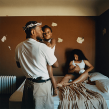 In a bedroom, Kendrick Lamar wearing woven crown of thorns is seen holding an infant child with a woman seen sitting on a bed holding another baby. The bedroom has brown paint with white spots and a radiator.
