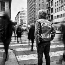 Jon Bon Jovi on a city street with his back turned to the camera, wearing a jacket that has the band name and album title on it
