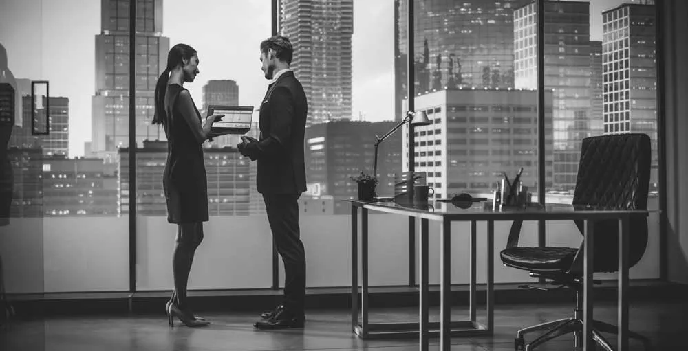 A man and a woman standing by the window and looking at a laptop near the office desk and chair