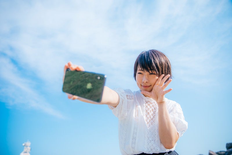 青空の下でスマホで自撮りする女子の写真