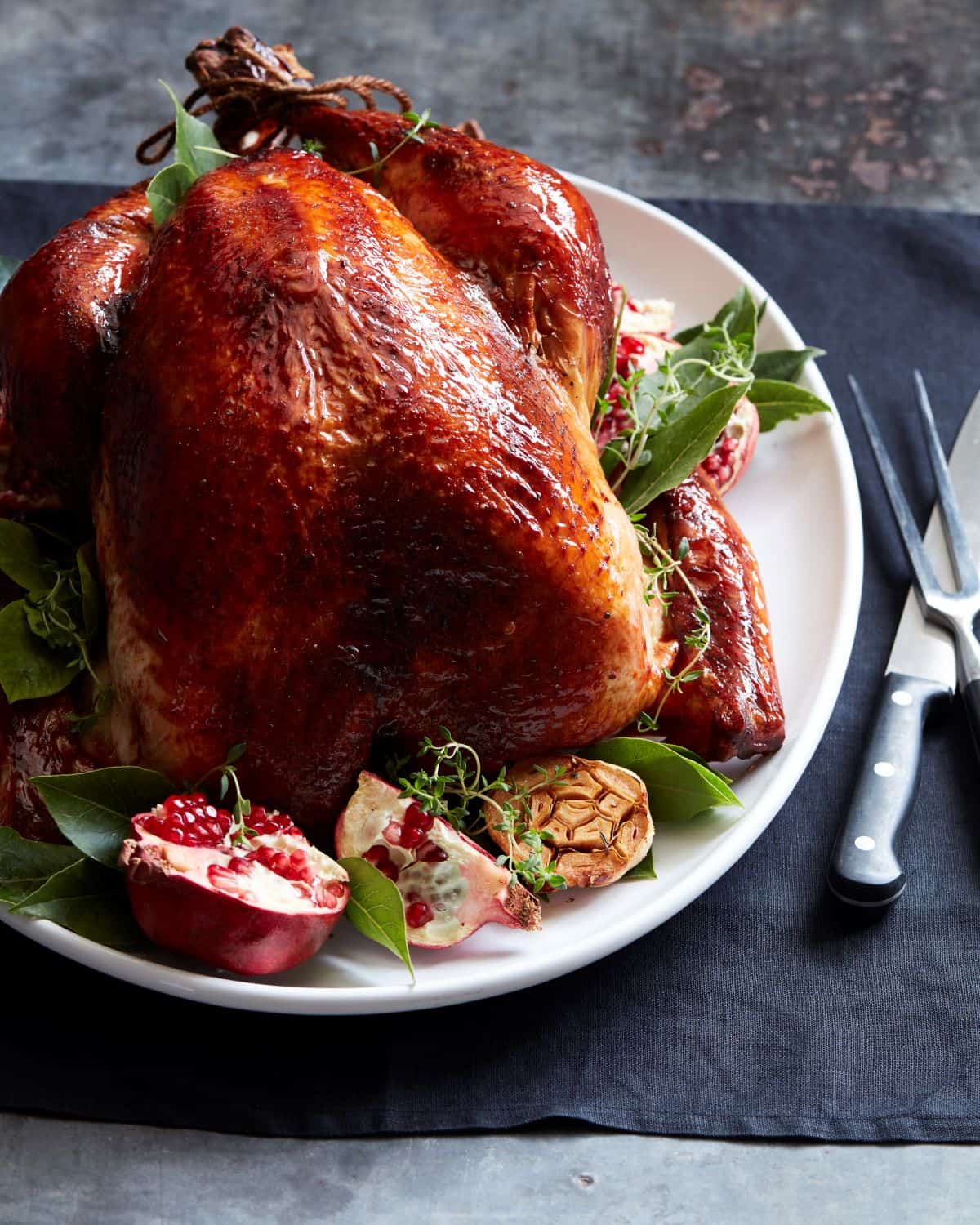 A white plate with a roasted turkey with pomegranates, halved garlics and fresh herbs around it in the plate, and carving fork and knife by its side.