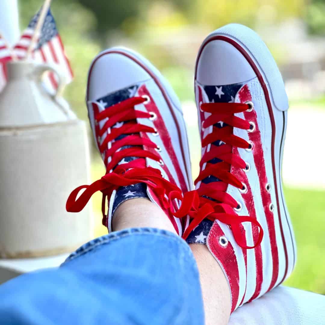 Person wearing red, white, and blue sneakers with star and stripe patterns. The shoes are shown prominently, with the person's legs crossed and a blurred background containing a white vase with small American flags.