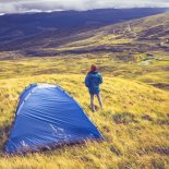 Article thumbnail: Woman is wild camping and standing next to her tent on a grassy mountain top