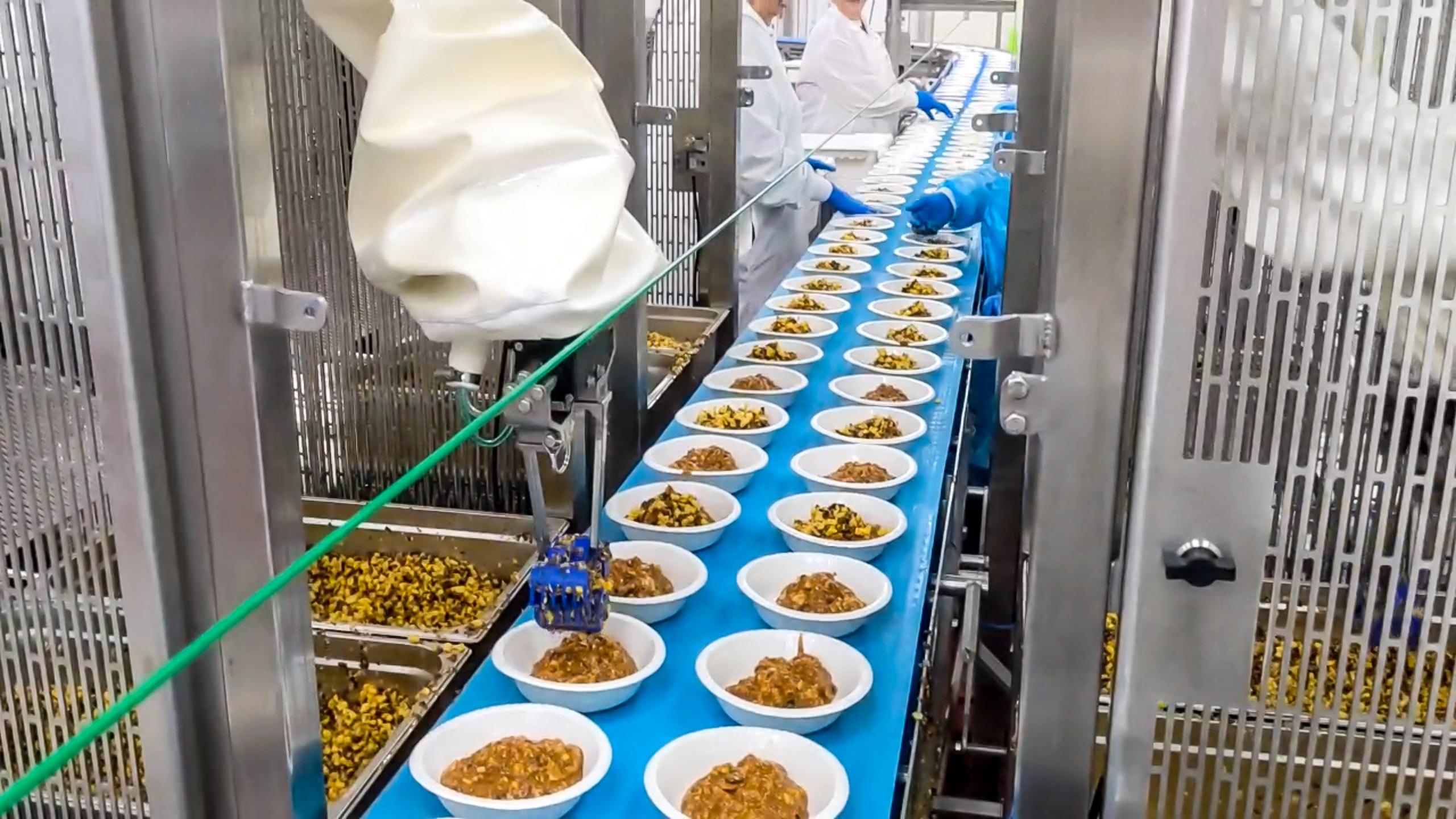 a conveyor with two rows of dishes. Human workers can be seen at stations behind a robot arm that is topping the bowls with a corn salad