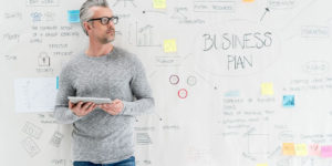 A man standing in front white wall adorned with writing and brainstorming ideas relating to a business plan.