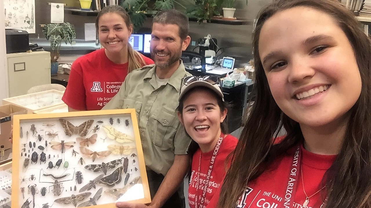 Three female students pose for a photo with an APHIS entomologist and his insect collection.