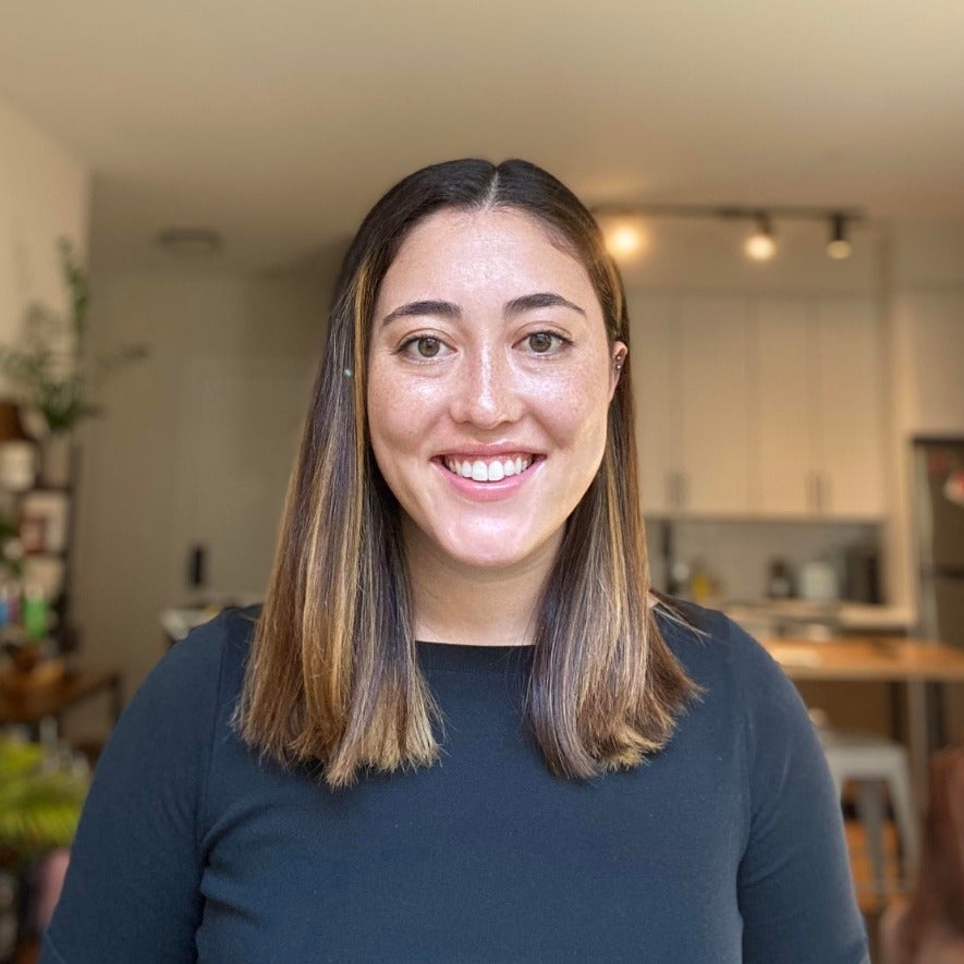 Isabella Sarmiento's headshot. A person with straight, medium length hair wearing a long-sleeved t-shirt is inside an apartment, smiling at the camera.