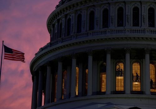 The very top of the US Capitol building is visible with a pink sunset in the background and a US flag on a flagpole to the side.