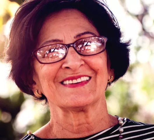 Imagem contém uma senhora de oculos de grau e blusa listrada preto e branco, sorrindo.