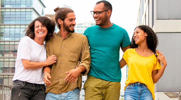 Imagem contém um grupo de quatro pessoas felizes, no fundo da imagem prédios e céu claro.