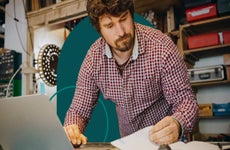 Man reviewing paperwork in front of a laptop in his workshop.