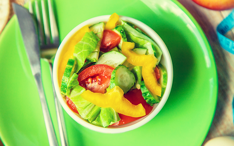 Imagem da publicação: "Como melhorar a memória" representando o cuidado com a alimentação. Prato verde com talheres ao lado de pote com uma salada de alface, pimentão amarelo, tomates cortados e alface. 