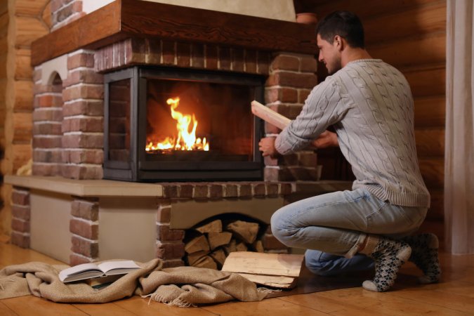 Man putting dry firewood into brick-surrounded fireplace at home.