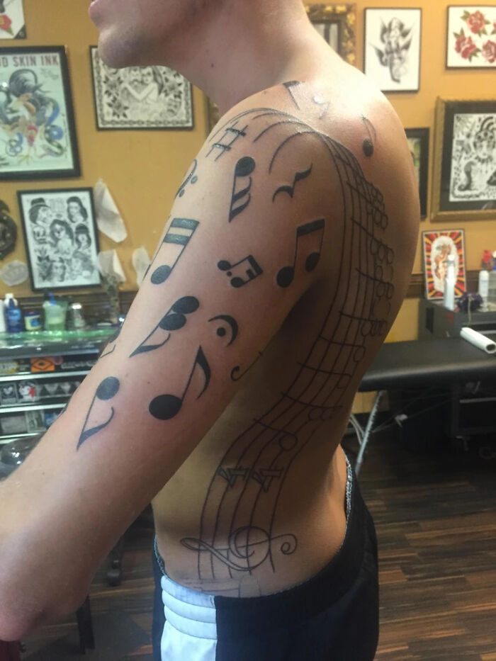 Music tattoo featuring musical notes flows from shoulder to arm in a tattoo studio setting.