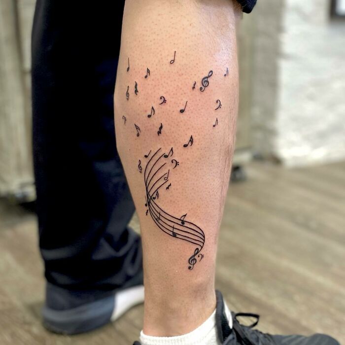 Music tattoo of notes and staff on a person's leg, featuring intricate design with floating musical symbols.