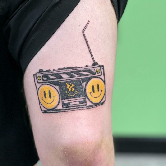 Tattoo of a retro boombox with yellow smiley faces, symbolizing music on an upper arm with a green background.