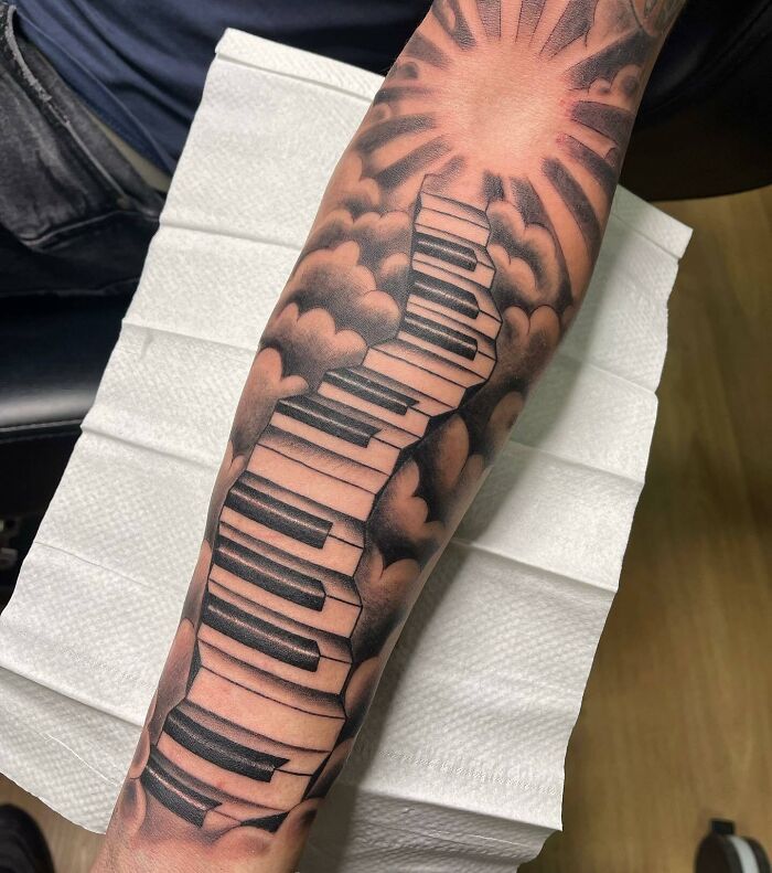 Tattoo of a piano keyboard winding through clouds to a sunburst, representing a music theme on an arm.