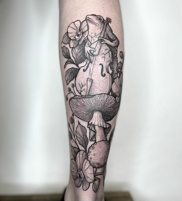 Frog playing cello in intricate music tattoo design on leg.