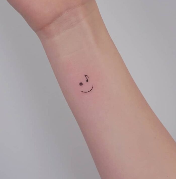 Minimalist music tattoo on forearm with musical note and asterisk, forming a smiley face design.