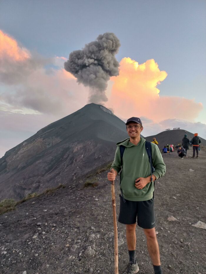 A person stands with a hiking stick near an erupting volcano, showcasing incredible world photos.